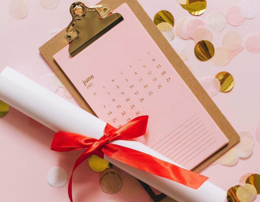 A rolled diploma with a calendar and confetti on a pink background symbolizing graduation.